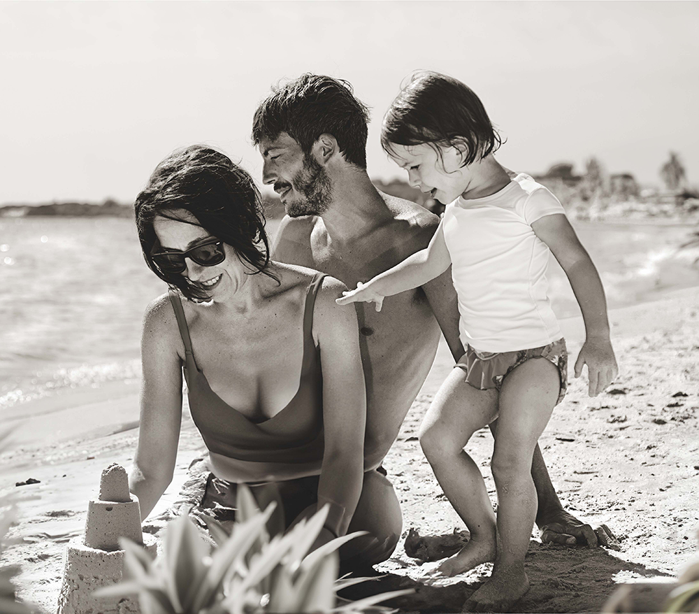 Una famiglia composta da madre, padre e figlia gioca sulla spiaggia, impegnata a costruire castelli di sabbia. La madre sorride mentre aggiunge dettagli al castello, il padre la osserva sorridente e la bambina, curiosa, tende la mano verso la sabbia. L’immagine in bianco e nero e l’atmosfera rilassata evocano un momento di affetto e complicità familiare in riva al mare. Fotografia di ritratto professionale che cattura un ricordo intimo e felice.