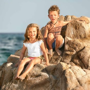 Due bambini sorridenti ripresi frontalmente, seduti su grandi scogli di fronte al mare. La bambina indossa una maglietta bianca e costume colorato, mentre il bambino indossa una maglietta beige con disegno e costume. Entrambi hanno i piedi coperti di sabbia e appaiono rilassati e felici sotto la luce calda del sole. Sullo sfondo, l’acqua blu e l’orizzonte aperto completano la scena, trasmettendo un senso di libertà e gioia estiva. Fotografia di ritratto spontaneo e naturale in un contesto marino.